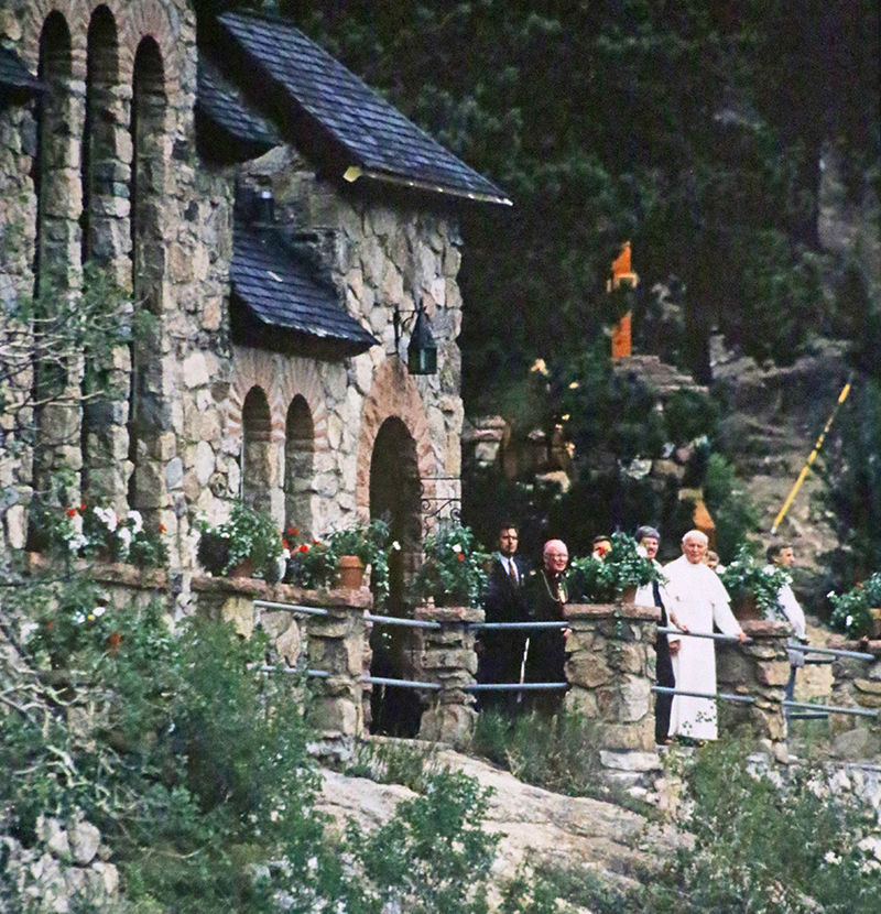 A stone chapel with steep shingled roofs and a cross stands on a rock outcrop above a grassy pond, framed by pines.