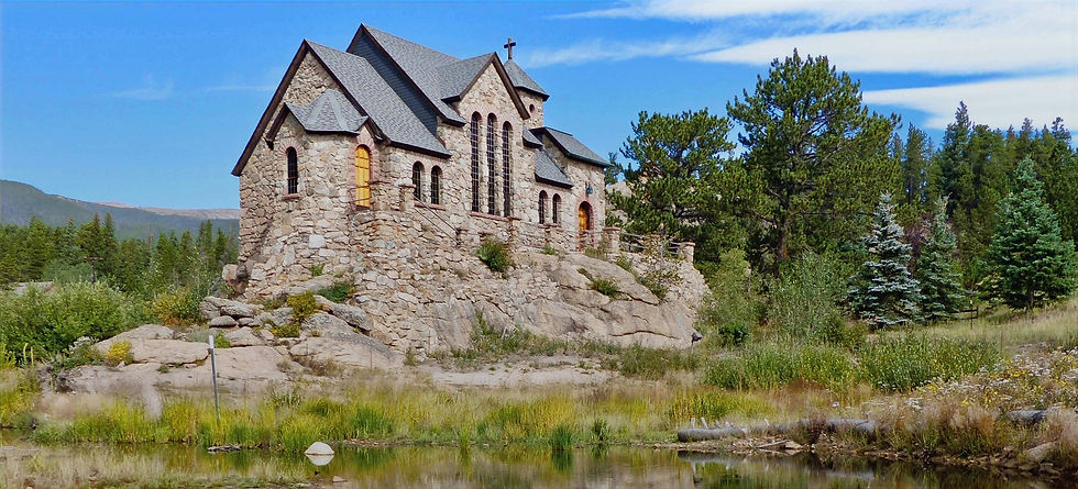 A stone chapel with steep shingled roofs and a cross stands on a rock outcrop above a grassy pond, framed by pines.