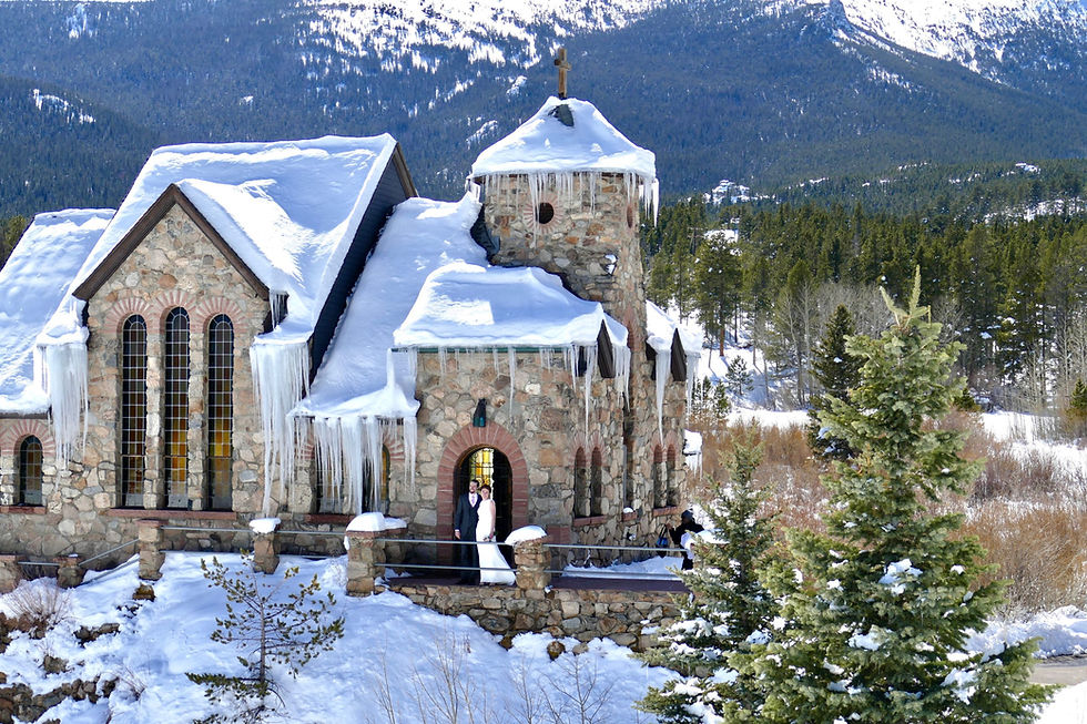 A stone chapel with steep shingled roofs and a cross stands on a rock outcrop above a grassy pond, framed by pines.